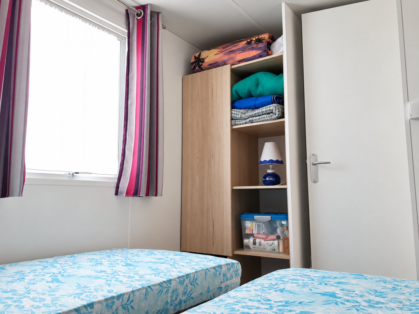 Chambre avec deux lits, une armoire et des rideaux rayés devant la fenêtre.