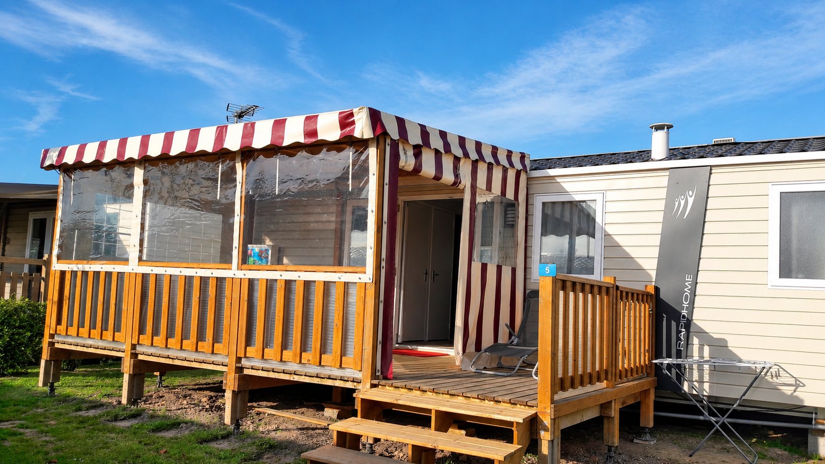 Mobil-home avec terrasse en bois couverte et store rayé rouge et blanc.