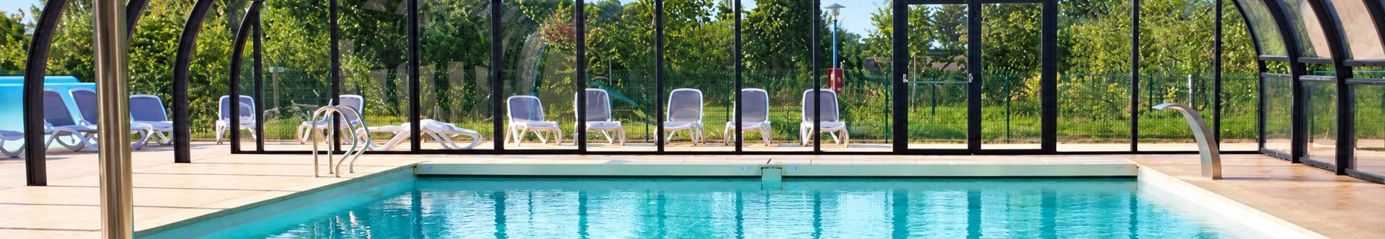 La piscine couverte du camping sous son abri vitré, avec des chaises longues et de la verdure.