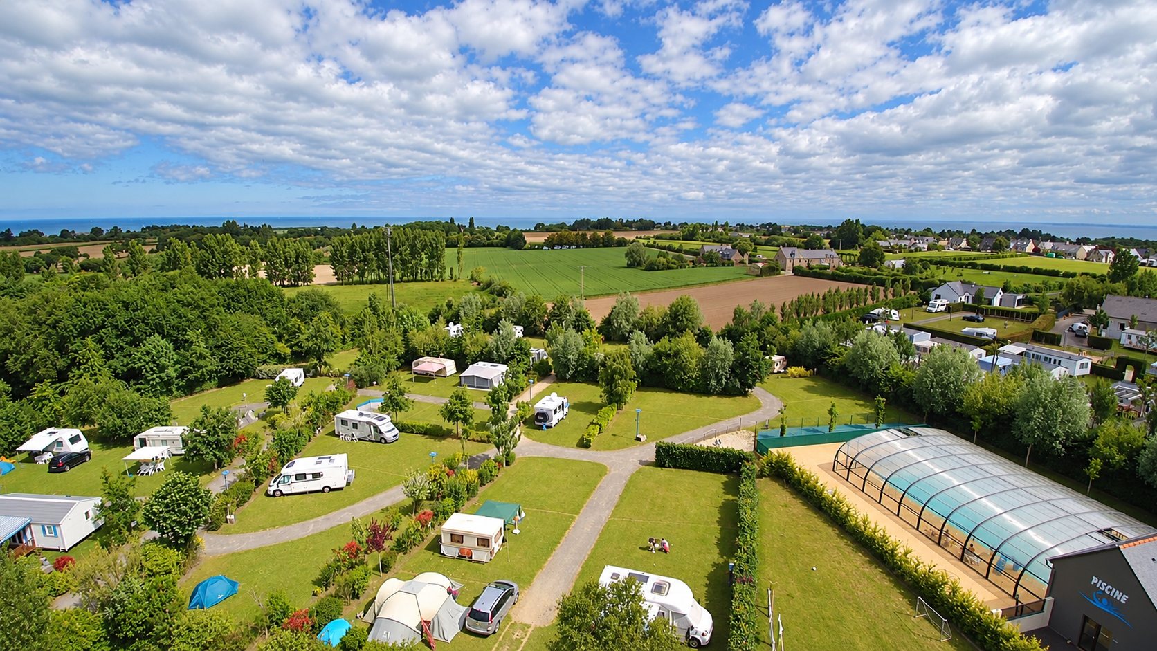 Vue aérienne du camping Les Clos Fleuris : emplacements dans la verdure, caravanes, piscine couverte et la mer à l’horizon.